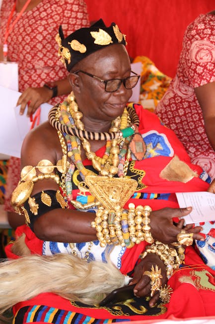 This image features a woman dressed in traditional African attire, including a vividly patterned red garment and an elaborate array of gold and beaded jewelry, such as necklaces, bracelets, and headpi